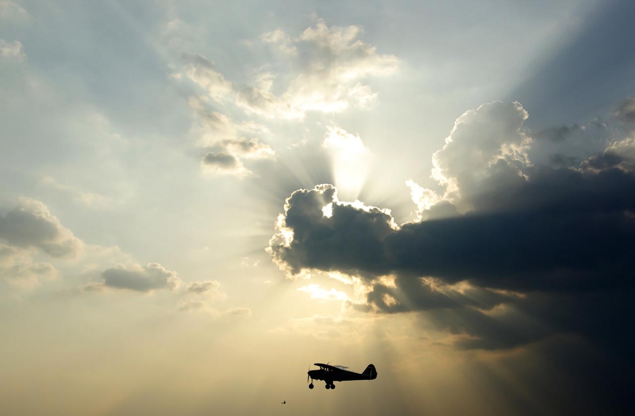 Silhouette of a small single-engine airplane flying beneath dramatic clouds as sunlight breaks through the sky.